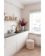 Et moderne, minimalistisk køkken med hvide skabe, en sort bordplade og en kunstig hortensiablomst 55 cm mørk pink i en vase nær bordpladen, plus træhylder med indretning og en rund skammel med hynder nær et vindue med persienner.