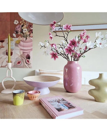 Et moderne rum har et dækket bord med kunstig gren Magnolia lille blomstret 89 cm pink i en pink vase, pastelfarvede lysestager, skåle, kopper, en grøn skulpturel vase, en pink bog og blomsterkunst på væggen.