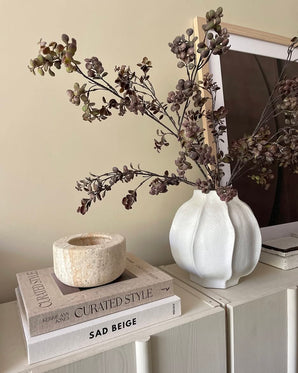 På et lysskab er der en hvid vase fyldt med den kunstige gren Eucalyptus bær 110 cm brun, ved siden af ​​en stak bøger og en sten lysestage, med et indrammet foto i baggrunden.