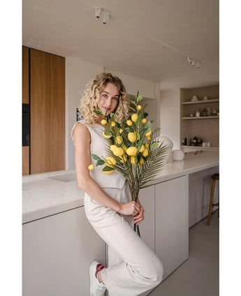 En kvinde med krøllet blond hår, klædt i et cremefarvet outfit, læner sig op ad en disk og holder buketten 'Artificial Branch Lemon 104 cm yellow', dekoreret med frodigt grønt. Det moderne køkken er indrettet i neutrale farver og har træaccenter.