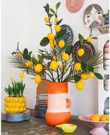 Et lyst rum med en høj vase fuld af citroner og grønne blade, hvor den markante Kunsttak Lemon 104 cm gul indtager en fremtrædende plads i arrangementet, omgivet af farverige keramikskåle, en skål med citroner og dekorative tallerkener på væggen - hvilket skaber et muntert, eklektisk udtryk.
