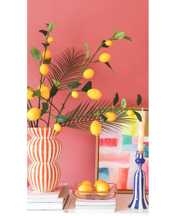 Mod en koralrosa væg står en stribet vase fyldt med Kunsttak Citrongrene på 104 cm i gult, et farverigt maleri, en stak bøger, en blå lysestage med et lys og en bakke citroner.