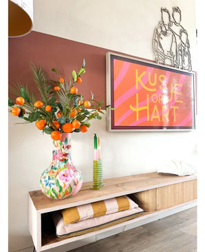 Op een houten plank staat een kleurrijke vaas met daarin de Kunsttak Mandarijn 93 cm oranje, naast een ingelijst kunstwerk met roze strepen waarop „Kusje op je Hart” staat. Op de plank liggen ook een groene kaars en opgevouwen dekens.