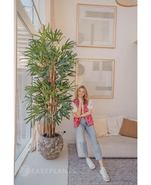 En kvinde i en pink mønstret cardigan og jeans smiler på en beige sofa ved siden af ​​Lady Artificial Palm 210 cm i en lys, moderne stue med abstrakt kunst på væggen.