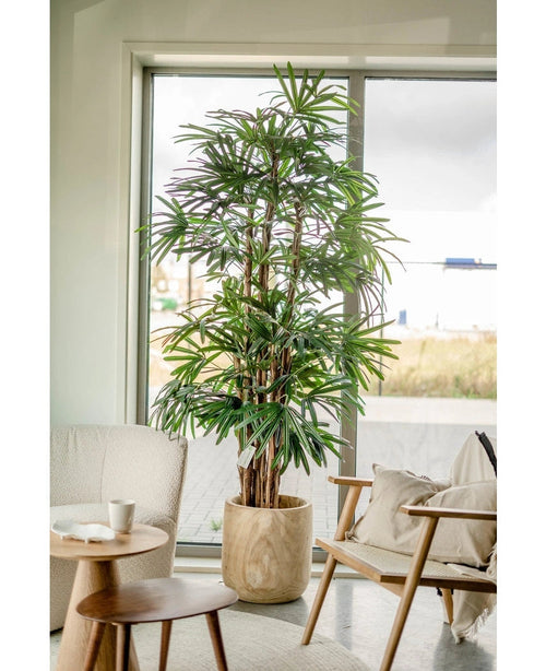 En 210 cm høj Lady Artificial Palm står ved siden af ​​et vindue i en lys, moderne stue med en polstret stol, et lille rundt bord og neutral indretning.