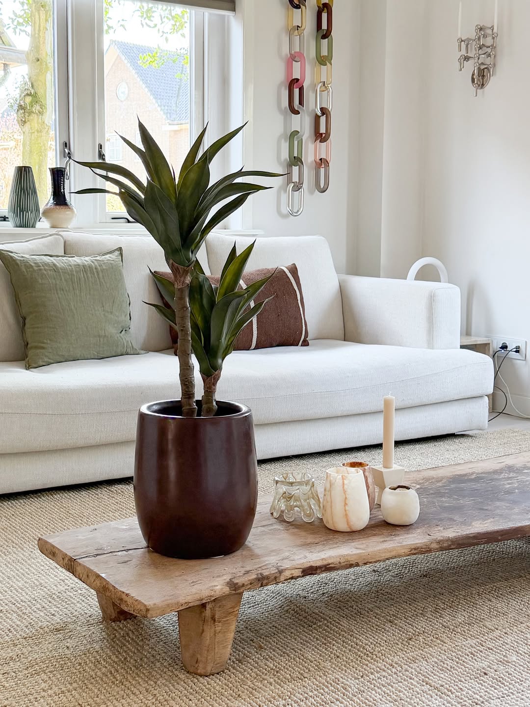 En moderne stue med en hvid sofa, grønne og brune hynder, et sofabord i træ dekoreret med stearinlys og den kunstige agaveplante 76 cm, og en vægdekoration i kædenet. Der kommer naturligt lys ind gennem vinduerne bag sofaen.