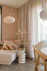 Et hyggeligt opholdsområde med en beige chaiselong, en stribet pude, den Artificial Caladium 90 cm pink, der giver et strejf af grønt, en moderne lampe, lyse gardiner og et trægulv. Blødt naturligt lys kommer ind gennem de gennemsigtige gardiner.